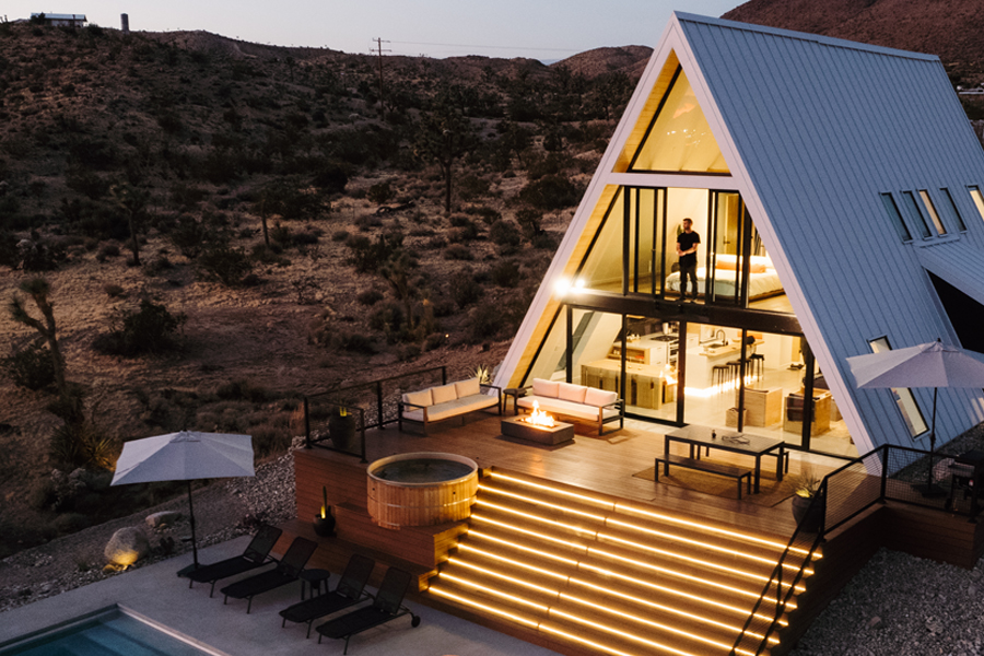 The Hata A-frame property in Joshua tree, photo taken at night. Showing the A-frame home, outdoor furniture on the deck, and chaise lounges by the pool.